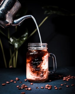 A metal container pours milk into a transparent mason jar filled with iced coffee. The swirling mix of milk and coffee creates a marbled effect. Coffee beans are scattered on a dark surface. In the background, there are blurred green leaves.