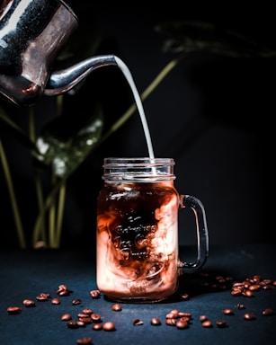 A metal container pours milk into a transparent mason jar filled with iced coffee. The swirling mix of milk and coffee creates a marbled effect. Coffee beans are scattered on a dark surface. In the background, there are blurred green leaves.