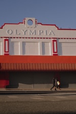 A large building with the word 'Olympia' displayed prominently at the top, featuring a red and white color scheme. The structure has decorative architectural elements and a closed storefront with a red awning. Two people walk on the sidewalk in front of the building, casting shadows on the ground in the afternoon light.