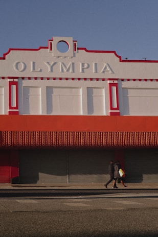 A large building with the word 'Olympia' displayed prominently at the top, featuring a red and white color scheme. The structure has decorative architectural elements and a closed storefront with a red awning. Two people walk on the sidewalk in front of the building, casting shadows on the ground in the afternoon light.