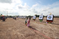 A series of archery targets set up on a large open field, each equipped with digital scoreboards showing zeros. A group of people stands on the left, under colorful umbrellas, possibly spectators or participants. Two individuals dressed in traditional clothing walk towards the targets in the foreground. The sky is partly cloudy, adding to the outdoor, competitive atmosphere.