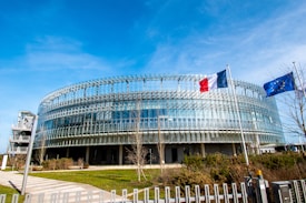 A modern circular glass building surrounded by landscaped greenery, with two flags mounted on flagpoles in front. The structure has a futuristic design with horizontal and vertical metal elements on the facade. A pathway leads towards the entrance of the building, and the sky is clear with bright blue tones.