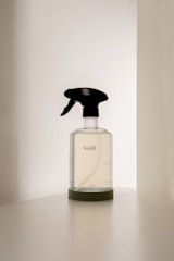 A transparent spray bottle with a black trigger nozzle rests on a solid surface. The bottle is labeled with the brand name 'kinfill' and contains a clear liquid. The background is minimalist with soft lighting that casts gentle shadows on the surface.