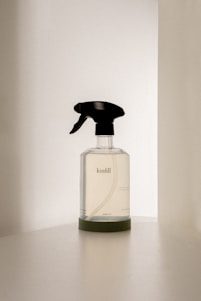 A transparent spray bottle with a black trigger nozzle rests on a solid surface. The bottle is labeled with the brand name 'kinfill' and contains a clear liquid. The background is minimalist with soft lighting that casts gentle shadows on the surface.