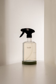 A transparent spray bottle with a black trigger nozzle rests on a solid surface. The bottle is labeled with the brand name 'kinfill' and contains a clear liquid. The background is minimalist with soft lighting that casts gentle shadows on the surface.