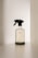 A transparent spray bottle with a black trigger nozzle rests on a solid surface. The bottle is labeled with the brand name 'kinfill' and contains a clear liquid. The background is minimalist with soft lighting that casts gentle shadows on the surface.
