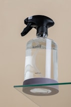 A clear spray bottle with a black trigger nozzle is placed on a glass surface. The bottle is transparent, partially filled with liquid, and has the word 'kinfill' printed on it.