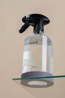 A clear spray bottle with a black trigger nozzle is placed on a glass surface. The bottle is transparent, partially filled with liquid, and has the word 'kinfill' printed on it.