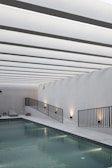 A sleek, modern indoor swimming pool area with minimalistic design. The ceiling features a series of evenly spaced linear white beams creating a rhythmic visual pattern. The pool is bordered by a linear walkway with black metal railings and ambient wall lighting. Modern lounge chairs and a small side table are situated on the poolside.