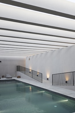 A sleek, modern indoor swimming pool area with minimalistic design. The ceiling features a series of evenly spaced linear white beams creating a rhythmic visual pattern. The pool is bordered by a linear walkway with black metal railings and ambient wall lighting. Modern lounge chairs and a small side table are situated on the poolside.
