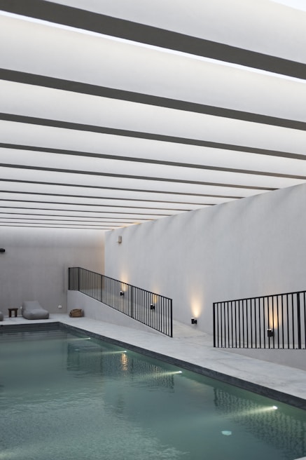 A sleek, modern indoor swimming pool area with minimalistic design. The ceiling features a series of evenly spaced linear white beams creating a rhythmic visual pattern. The pool is bordered by a linear walkway with black metal railings and ambient wall lighting. Modern lounge chairs and a small side table are situated on the poolside.