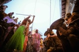 A lively street scene depicting a traditional festival with people dressed in colorful attire. A woman in a bright green and orange dress is holding a stick above her head, surrounded by others in vibrant garments. Some men are shielding themselves with large round objects, possibly participating in a cultural performance. The atmosphere is festive and dynamic, with onlookers gathered closely around.