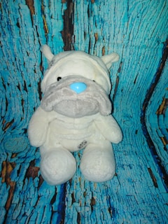 A plush toy of a dog with a white body, gray snout, and blue nose is placed against a backdrop resembling cracked, painted blue and brown wood.