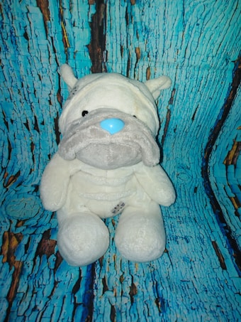 A plush toy of a dog with a white body, gray snout, and blue nose is placed against a backdrop resembling cracked, painted blue and brown wood.