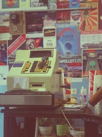 A vintage cash register with sunglasses resting on top is placed on a counter. The background is filled with colorful retro posters and advertisements. Below the register, there are some napkins, a Coca-Cola cup, and a few other small items.