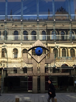 A modern glass building with the reflection of a historic architectural facade. Below the glass is a large artistic installation resembling an eye, positioned above a concrete structure with the text 'NOVA SCENA'. A person is walking past the building on a tiled pavement.