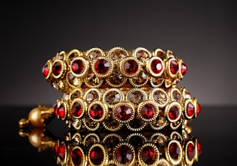 Ornate gold bracelets adorned with red and clear gemstones, arranged in a symmetrical pattern. The bracelets are highly reflective and rest on a glossy black surface, enhancing the luxurious feel.