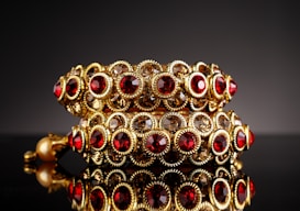 Ornate gold bracelets adorned with red and clear gemstones, arranged in a symmetrical pattern. The bracelets are highly reflective and rest on a glossy black surface, enhancing the luxurious feel.