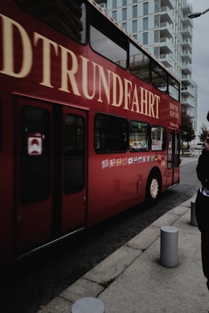 A red double-decker bus with the words 'Rundfahrt' and 'Hop On Hop Off' is on the street. It is parked near a modern building with multiple floors and glass windows. Cylindrical concrete bollards are visible on the sidewalk, and there is a person partially visible on the right side.
