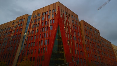 A modern architectural building with a striking red and tan facade dominates the scene. The structure features numerous rectangular windows arranged in a geometric pattern. A crane is visible against the overcast sky, indicating potential construction activity nearby.