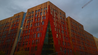 A modern architectural building with a striking red and tan facade dominates the scene. The structure features numerous rectangular windows arranged in a geometric pattern. A crane is visible against the overcast sky, indicating potential construction activity nearby.