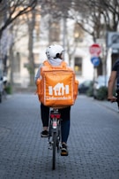 A person wearing a white helmet and casual clothing is riding a bicycle down a tree-lined street. They have a large orange delivery backpack with 'Lieferando.de' logo on it. The street is paved and lined with trees and blurred buildings in the background. A stop sign is visible on the right.