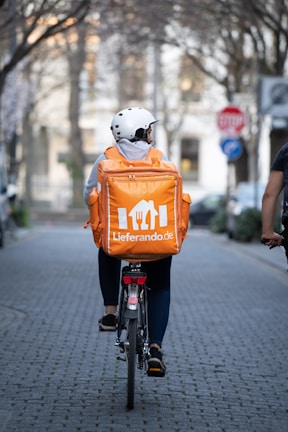 A person wearing a white helmet and casual clothing is riding a bicycle down a tree-lined street. They have a large orange delivery backpack with 'Lieferando.de' logo on it. The street is paved and lined with trees and blurred buildings in the background. A stop sign is visible on the right.
