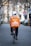 A person wearing a white helmet and casual clothing is riding a bicycle down a tree-lined street. They have a large orange delivery backpack with 'Lieferando.de' logo on it. The street is paved and lined with trees and blurred buildings in the background. A stop sign is visible on the right.