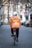 A person wearing a white helmet and casual clothing is riding a bicycle down a tree-lined street. They have a large orange delivery backpack with 'Lieferando.de' logo on it. The street is paved and lined with trees and blurred buildings in the background. A stop sign is visible on the right.