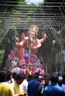 A large statue of a deity with multiple arms is being paraded through a street. The figure is intricately adorned with colorful garments and jewelry. Surrounding the statue are numerous people, some dressed in vibrant clothing, participating in the procession. Above the statue, decorative lights are strung across the street, and lush green trees line the path.