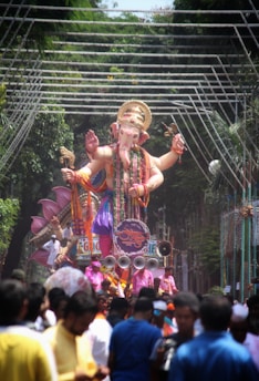 A large statue of a deity with multiple arms is being paraded through a street. The figure is intricately adorned with colorful garments and jewelry. Surrounding the statue are numerous people, some dressed in vibrant clothing, participating in the procession. Above the statue, decorative lights are strung across the street, and lush green trees line the path.