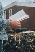 A person holds a book open in one hand and a rosary in the other. The background features greenery and an out-of-focus building. The person wears a sweater with a floral pattern.