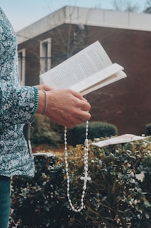 A person holds a book open in one hand and a rosary in the other. The background features greenery and an out-of-focus building. The person wears a sweater with a floral pattern.