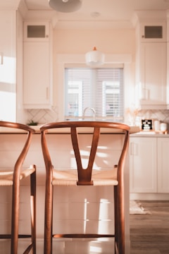 Two wooden bar stools with woven seats are positioned at a kitchen island in a light-filled, modern kitchen. The kitchen features white cabinets, a small window with horizontal blinds, and a sink with a faucet. Subtle decorations and a soft lighting give the space a cozy and inviting feel.