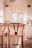 Two wooden bar stools with woven seats are positioned at a kitchen island in a light-filled, modern kitchen. The kitchen features white cabinets, a small window with horizontal blinds, and a sink with a faucet. Subtle decorations and a soft lighting give the space a cozy and inviting feel.
