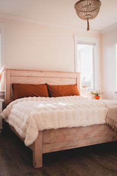 A cozy bedroom with a wooden bed frame and orange pillows. The bed is covered with a fluffy beige throw blanket. Natural light pours in through the windows, enhancing the room's warm tones. A small potted plant is placed by the window, and a woven light fixture hangs from the ceiling.