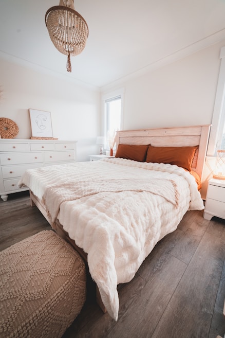 A cozy bedroom with a wooden bed frame and textured bedding, featuring a soft beige and brown color palette. A chandelier with intricate designs hangs from the ceiling. The room includes white dressers, a framed line art piece leaning on a dresser, and a lamp on a small side table. Natural light streams through a window, adding warmth to the space.