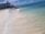 A serene beach scene featuring clear turquoise waters gently washing onto white sandy shores. In the distance, a large hotel building is visible along with a pier extending into the ocean. The sky is bright blue with a few scattered clouds.