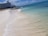 A serene beach scene featuring clear turquoise waters gently washing onto white sandy shores. In the distance, a large hotel building is visible along with a pier extending into the ocean. The sky is bright blue with a few scattered clouds.