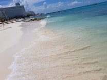 A serene beach scene featuring clear turquoise waters gently washing onto white sandy shores. In the distance, a large hotel building is visible along with a pier extending into the ocean. The sky is bright blue with a few scattered clouds.
