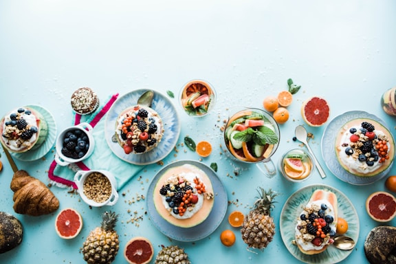 A vibrant and colorful spread of breakfast or brunch items including pancakes or waffles topped with various berries, cups of granola or seeds, slices of grapefruit and orange, a croissant, and a pitcher of water with slices of cucumber and mint leaves. Pineapple and other tropical fruit slices add an exotic touch.