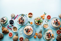 A vibrant and colorful spread of breakfast or brunch items including pancakes or waffles topped with various berries, cups of granola or seeds, slices of grapefruit and orange, a croissant, and a pitcher of water with slices of cucumber and mint leaves. Pineapple and other tropical fruit slices add an exotic touch.