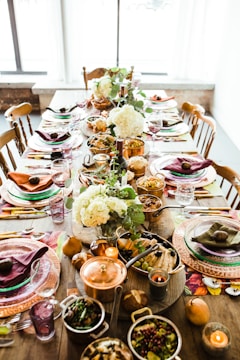 A dining table is elegantly set with plates, cutlery, glasses, and napkins arranged neatly. The centerpiece features white hydrangea flowers and green foliage. Various dishes are served in copper and glass containers, including fruit, vegetables, and bread. Wine glasses accompany each place setting, creating a sophisticated ambiance.