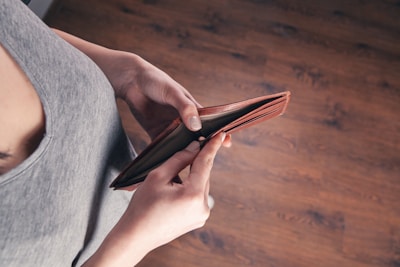 A person holding an empty brown wallet with both hands, viewed from above. The person is wearing a gray sleeveless shirt, and the background is a wooden floor.