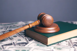 A wooden gavel resting on a green book surrounded by scattered dollar bills