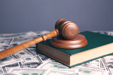 A wooden gavel resting on a green book surrounded by scattered dollar bills