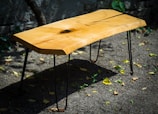A wooden table with a natural finish and a unique, rustic design. The tabletop features a distinctive wood grain pattern with a knot near the center. The table is supported by slender, black metal legs and is placed on a gravel surface outdoors. Some scattered leaves can be seen on the ground, creating a natural and organic setting.