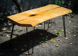 A wooden table with a natural finish and a unique, rustic design. The tabletop features a distinctive wood grain pattern with a knot near the center. The table is supported by slender, black metal legs and is placed on a gravel surface outdoors. Some scattered leaves can be seen on the ground, creating a natural and organic setting.