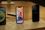 Several iPhones are displayed on a wooden surface. The central iPhone has its screen on, showing various app icons. Nearby, another iPhone has its screen off but is labeled 'iPhone 12 Pro Max.' The setup is organized on a display board with labels and charging cables.