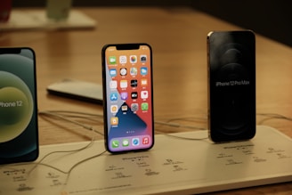 Several iPhones are displayed on a wooden surface. The central iPhone has its screen on, showing various app icons. Nearby, another iPhone has its screen off but is labeled 'iPhone 12 Pro Max.' The setup is organized on a display board with labels and charging cables.